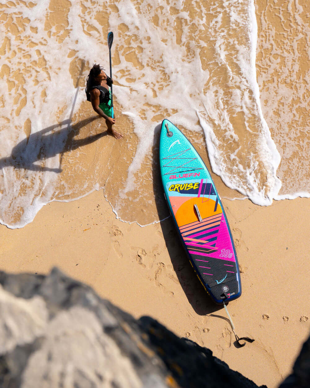 Paddleboard on a sandy beach with a person holding a paddle, waves lapping at the shore.