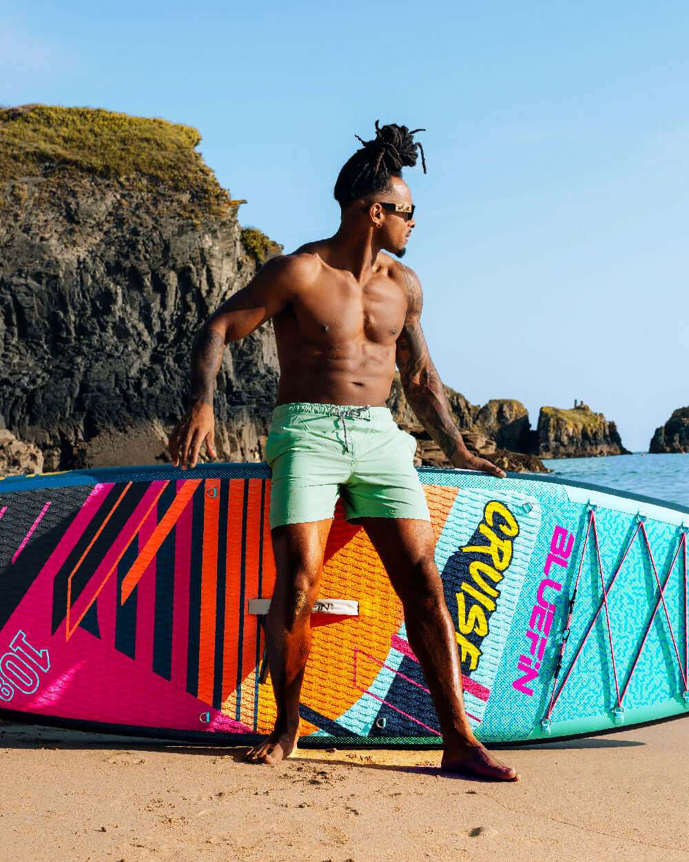 Man in swim shorts standing on a beach with a colorful Bluefin SUP paddle board. Rocky cliffs in the background.