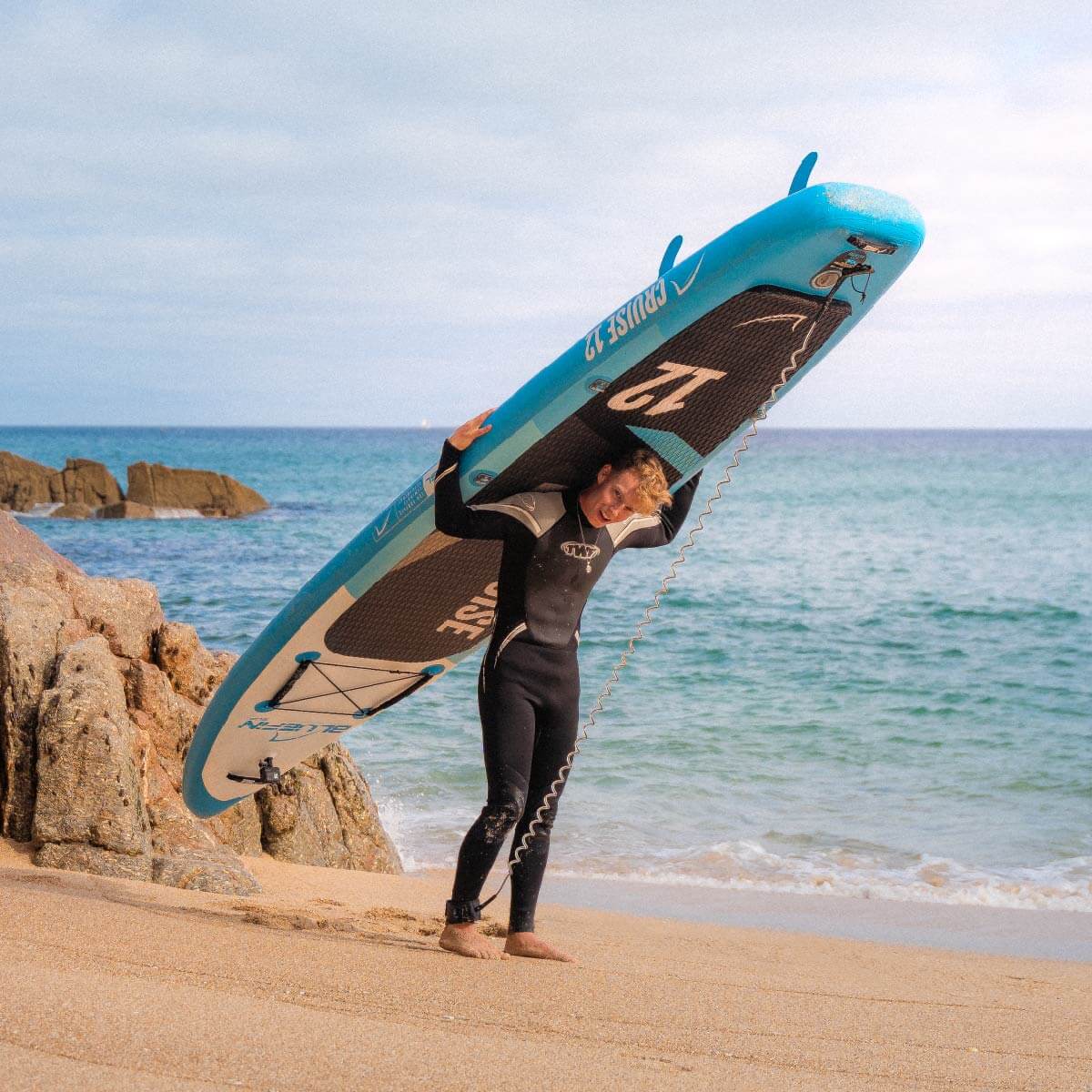 Person in a wetsuit carrying a blue paddleboard on a sandy beach with ocean waves in the background.