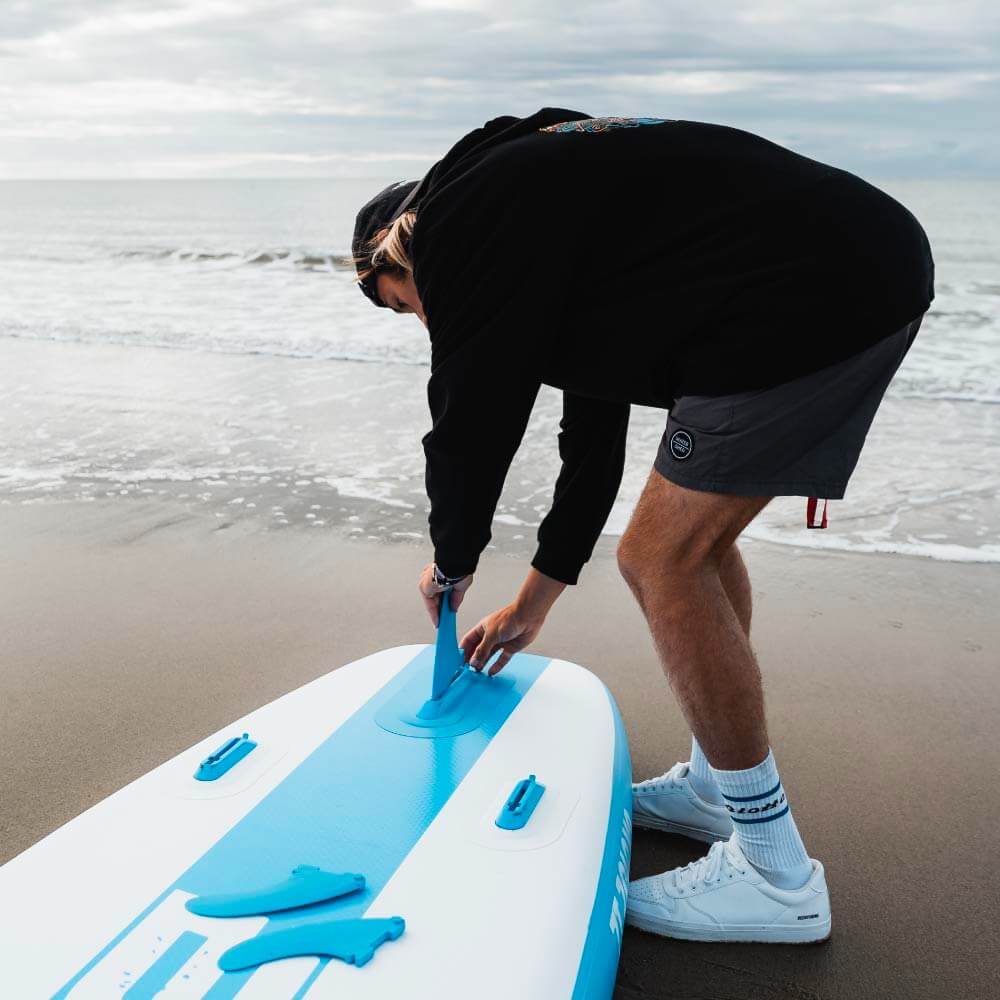 Person assembling a Bluefin paddleboard on the beach with waves in the background.
