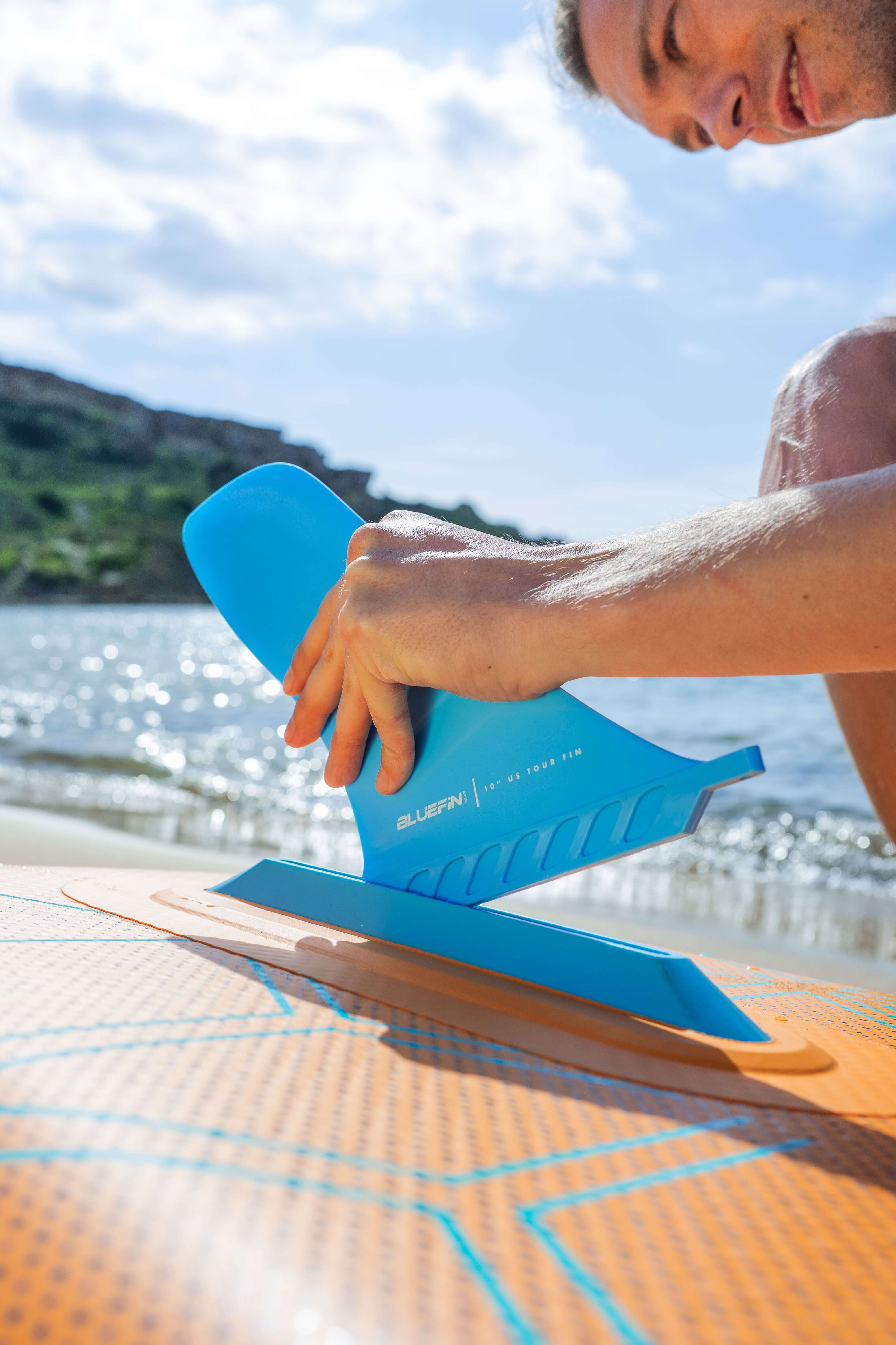 Person attaching a fin on a Bluefin cruise inflatable paddle board on a beach with water and sky in the background