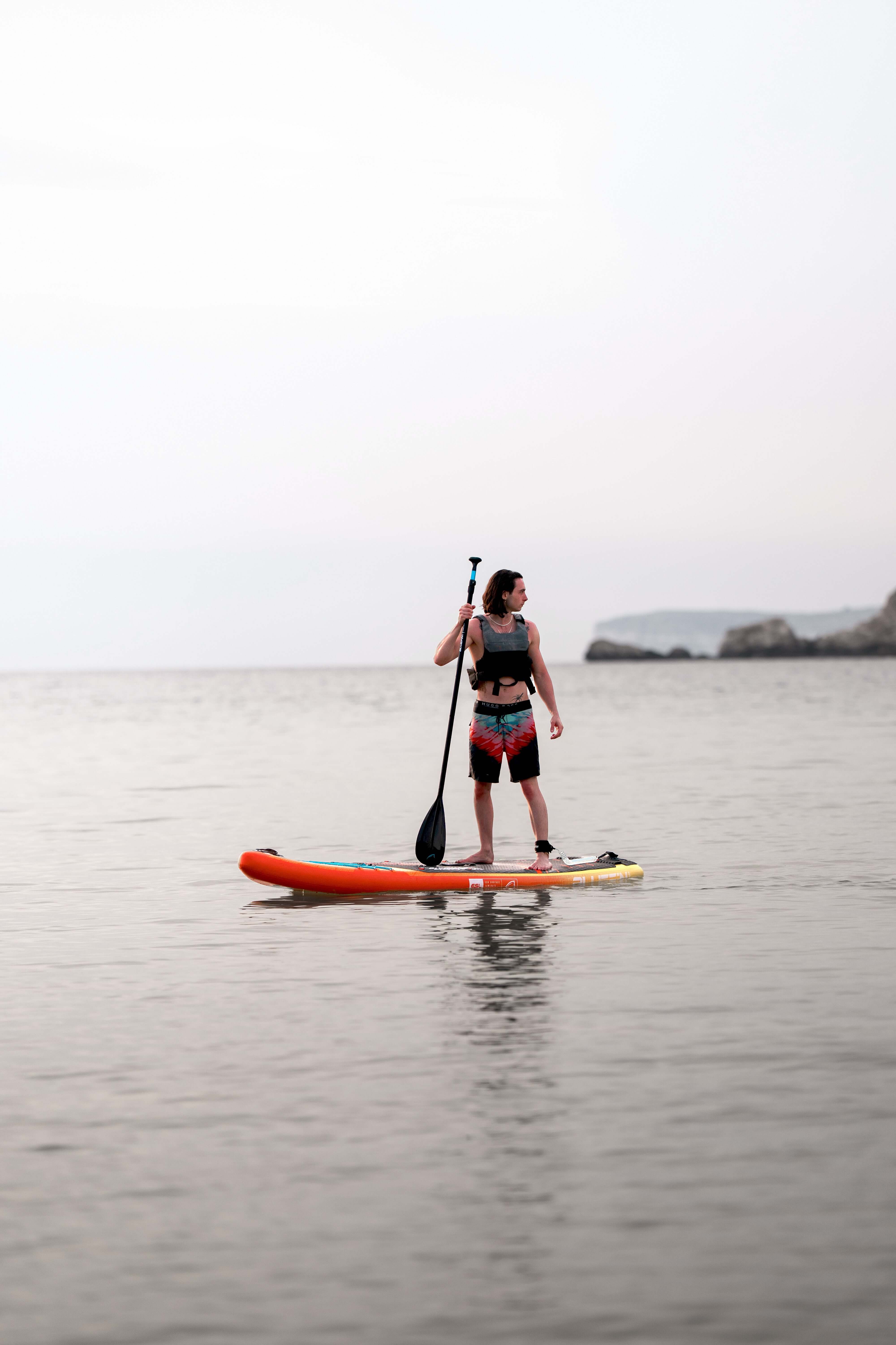 Paddleboarder standing on an orange SUP in calm water, looking towards the shore at sunset.