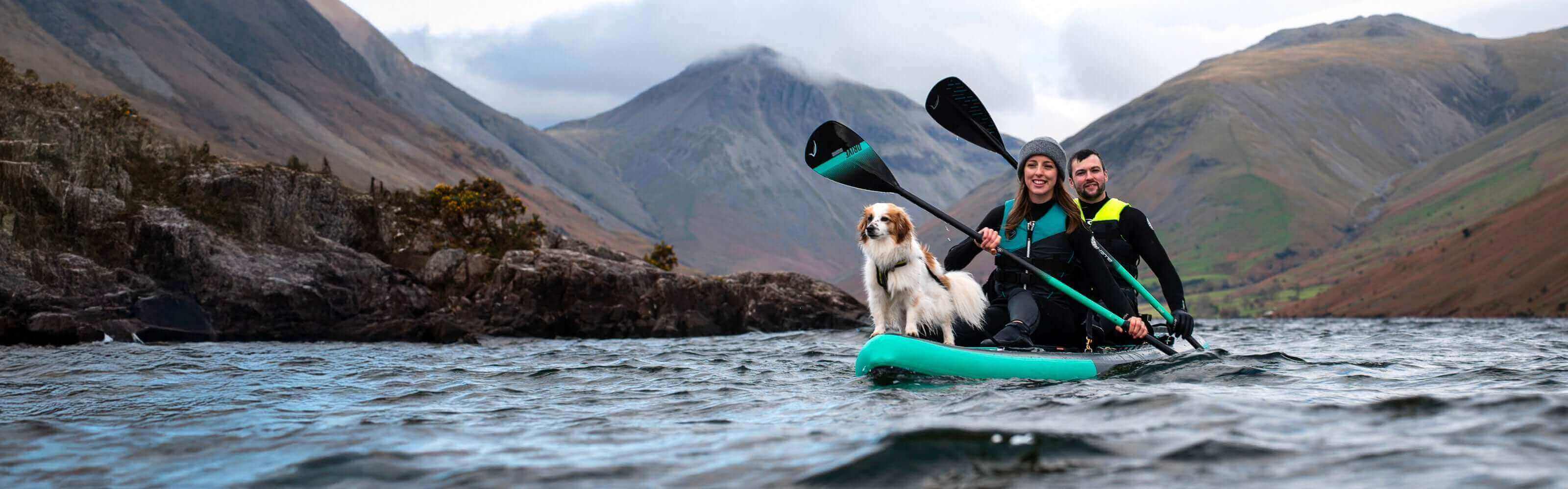 Two people and a dog on a teal inflatable paddleboard in a lake surrounded by mountains.