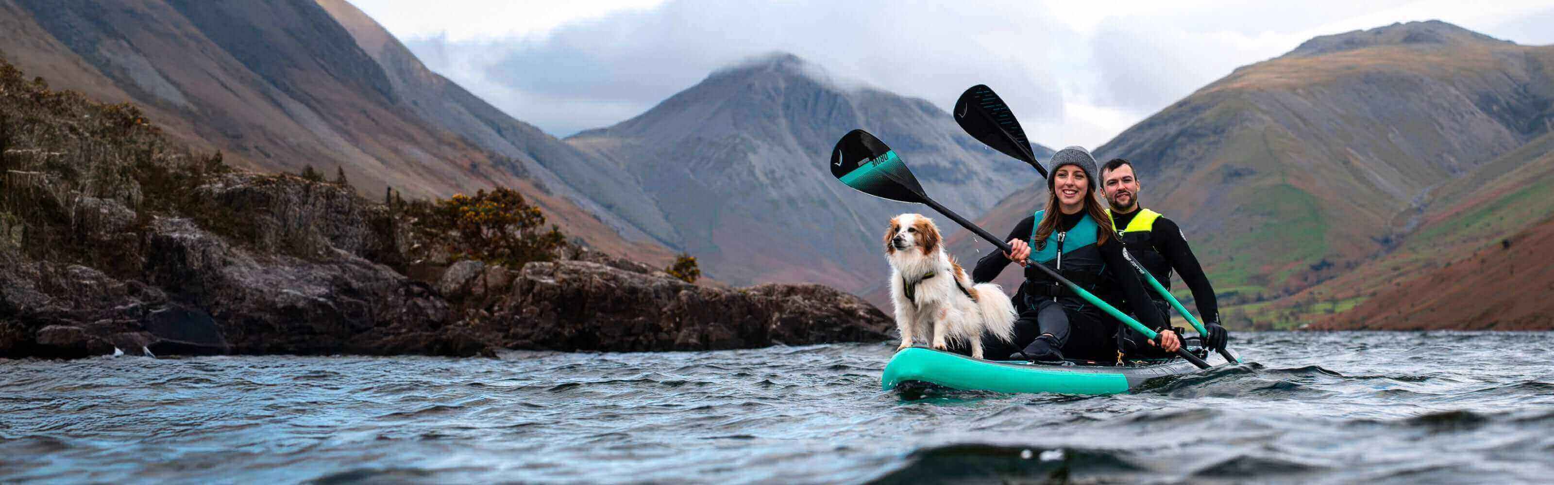 Two people and a dog on a teal inflatable paddleboard in a lake surrounded by mountains.