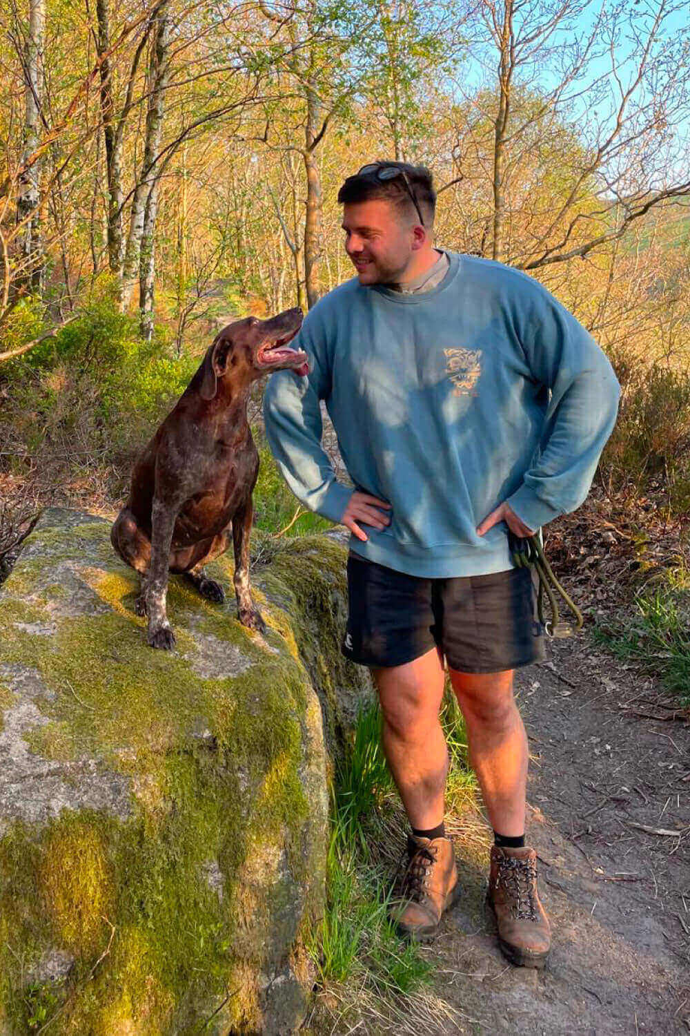 Man in a blue sweatshirt and shorts standing beside a brown dog on a mossy rock in a forest.