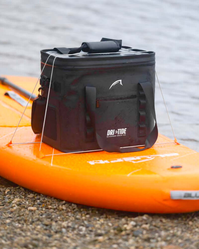 Black Dri-Tide cooler bag on an orange inflatable paddle board by a body of water.