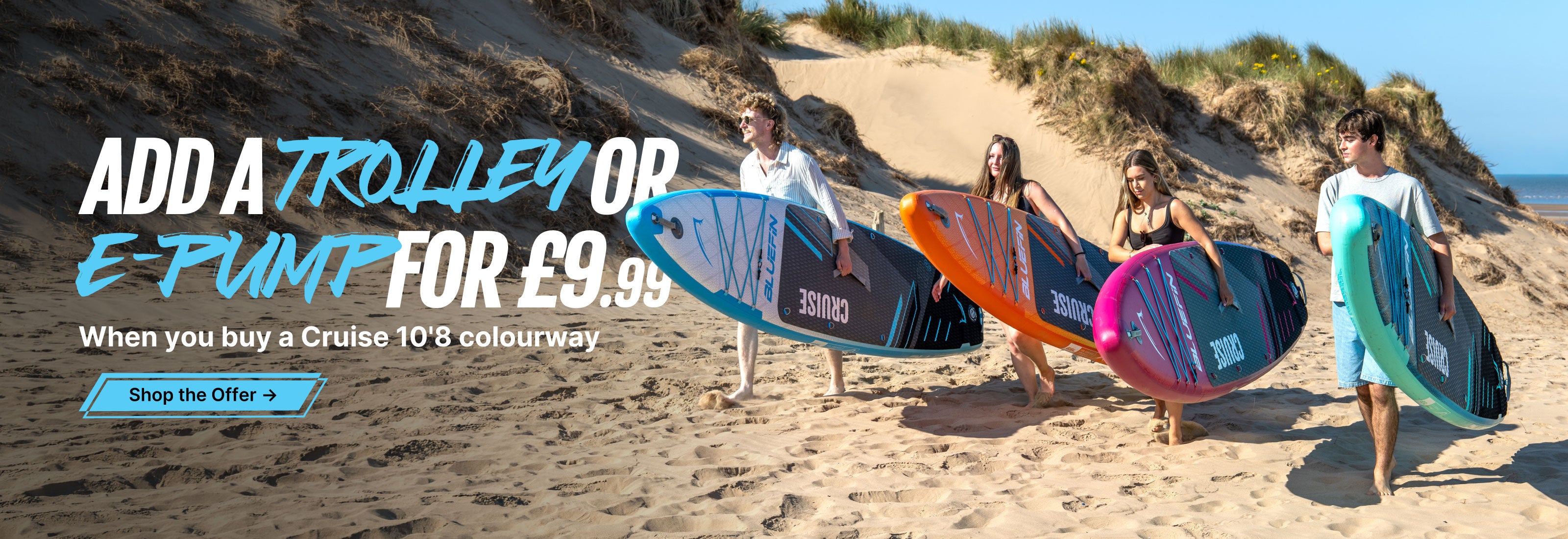 Group of four people carrying colorful stand-up paddleboards on a sandy beach with dunes in the background.