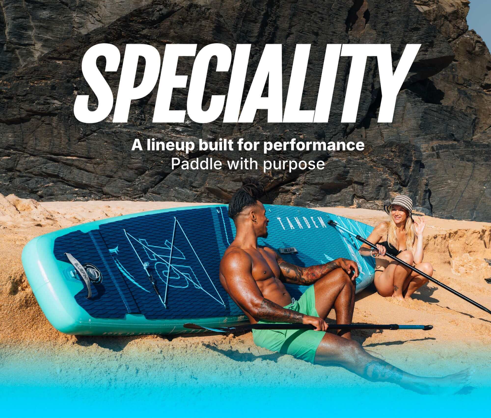 Two people on a sandy beach with a blue inflatable paddleboard and paddles, rocky background.