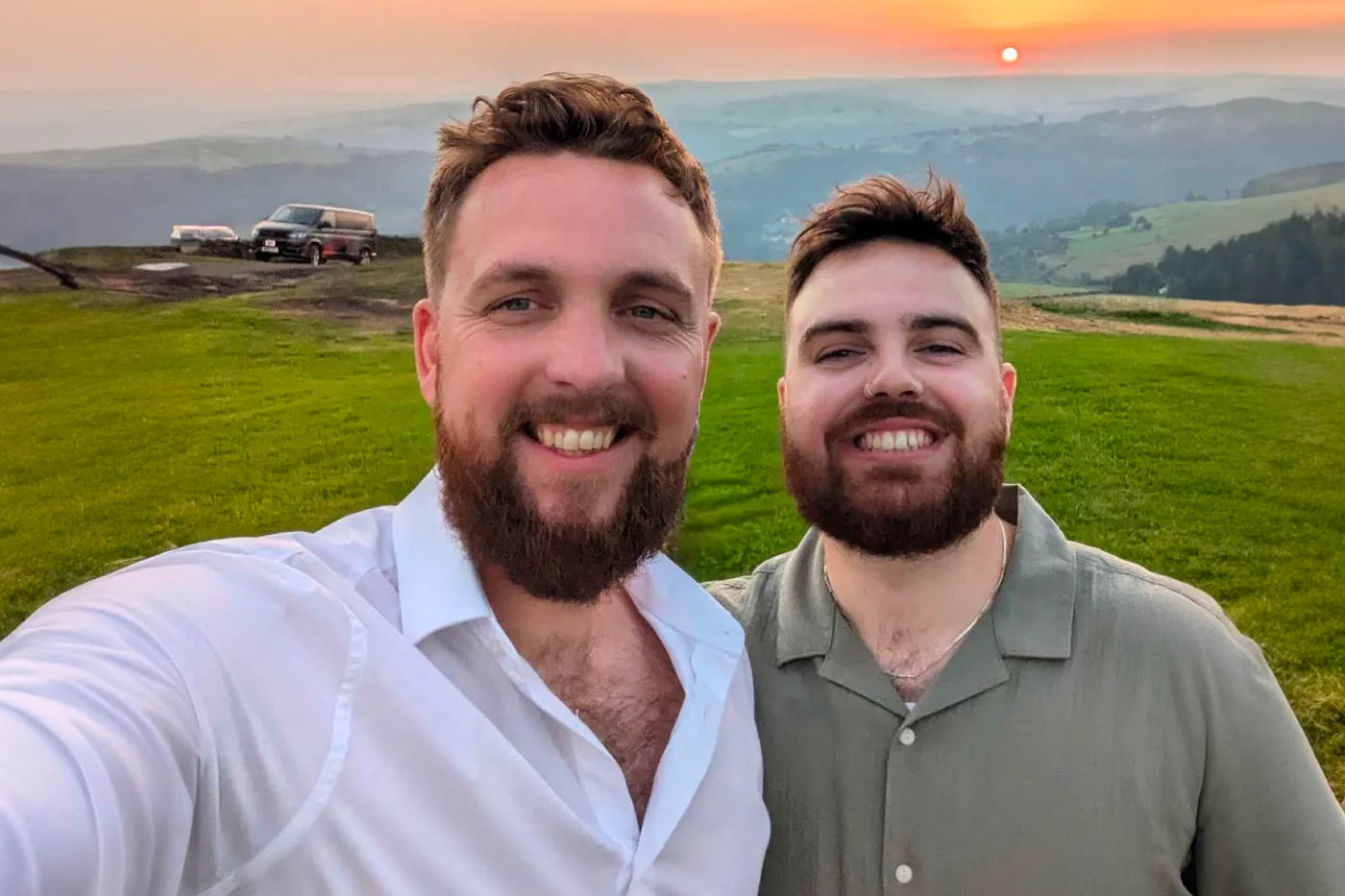 Two smiling men take a selfie against a scenic sunset backdrop with rolling hills and green grass.
