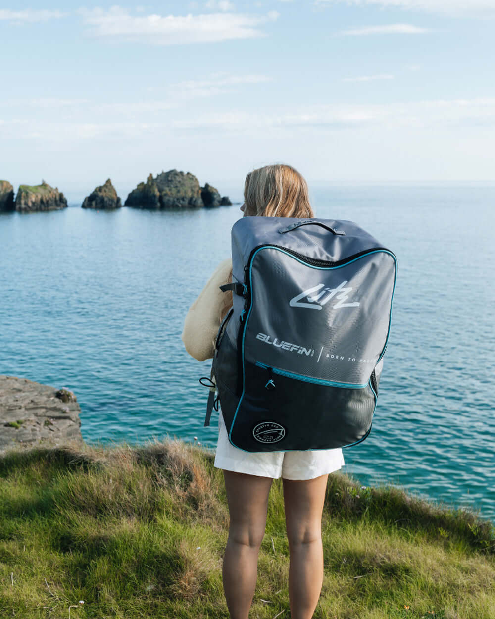 Person standing on grass by the water, wearing a backpack with Bluefin logo and gray color.