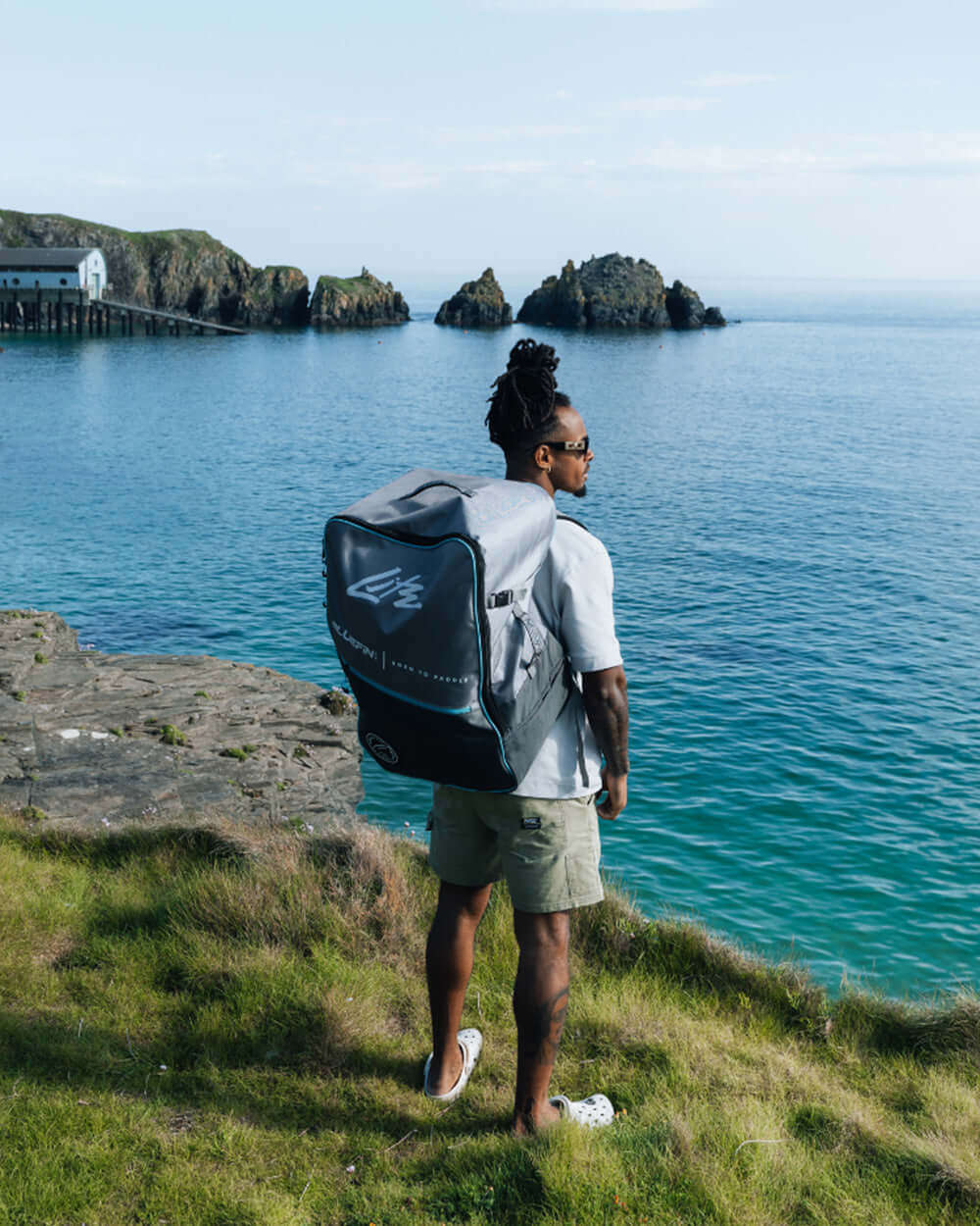 Person standing on grass by the sea, wearing a backpack and shorts, looking at rocky coastline.