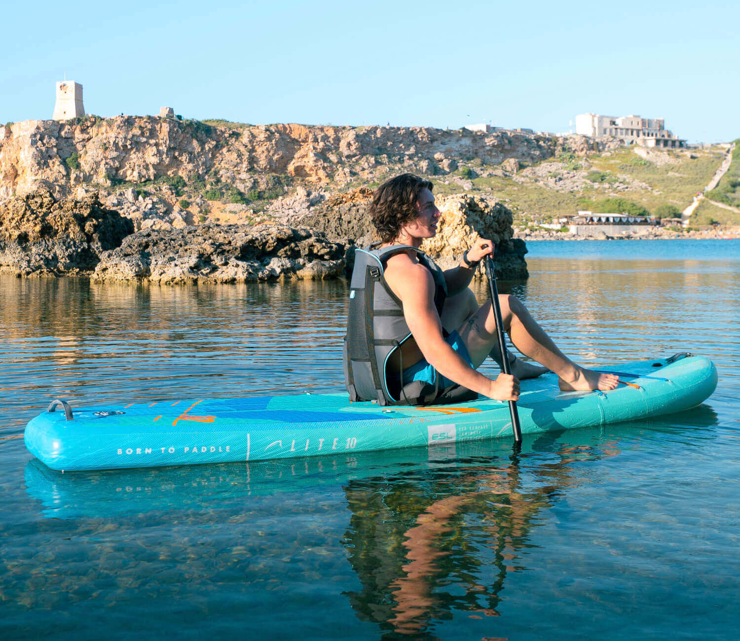 Person paddling on a turquoise inflatable paddleboard in calm water with rocky shoreline in background