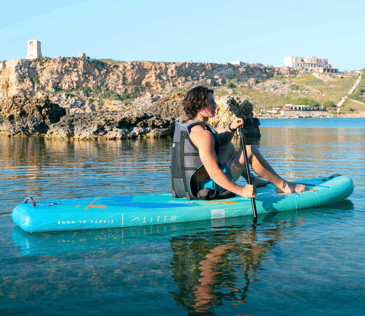 Person paddling on a turquoise inflatable paddleboard in calm water with rocky shoreline in background