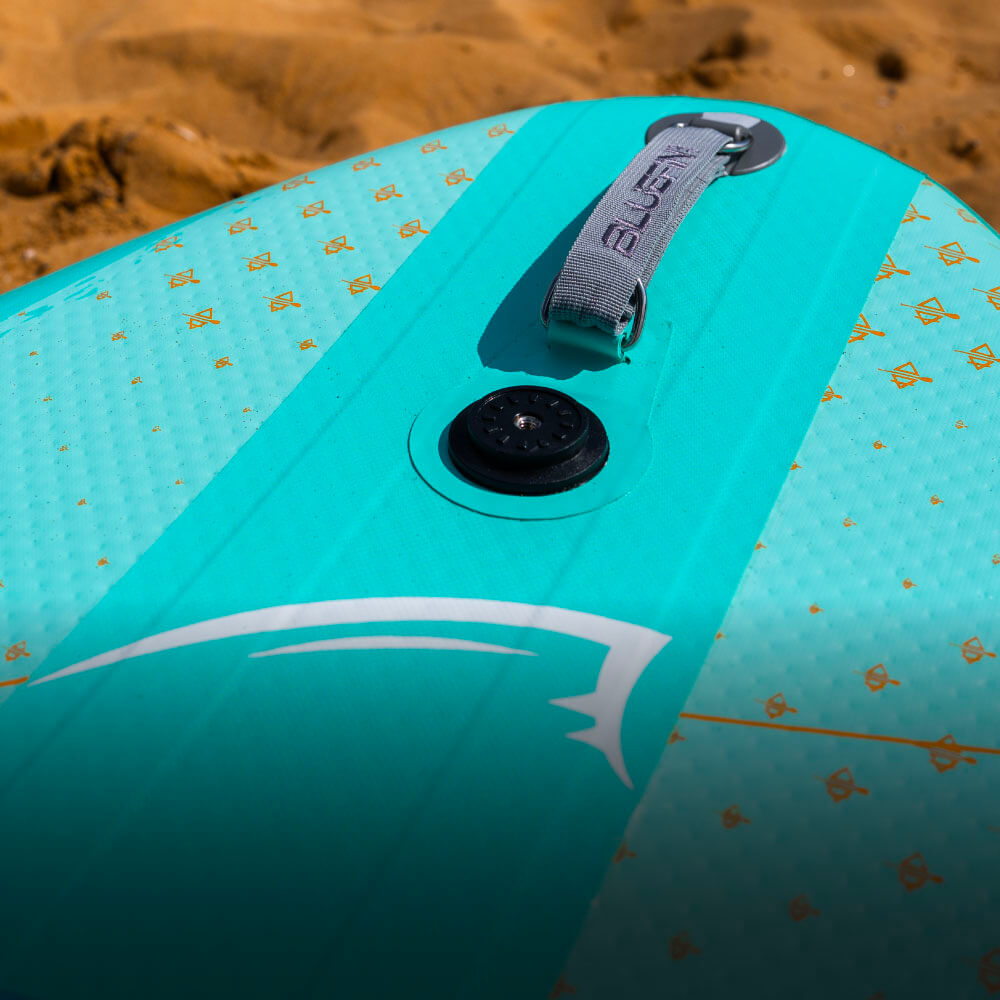 Close-up of a turquoise SUP with a grip handle and inflation valve, set against sandy beach backdrop.