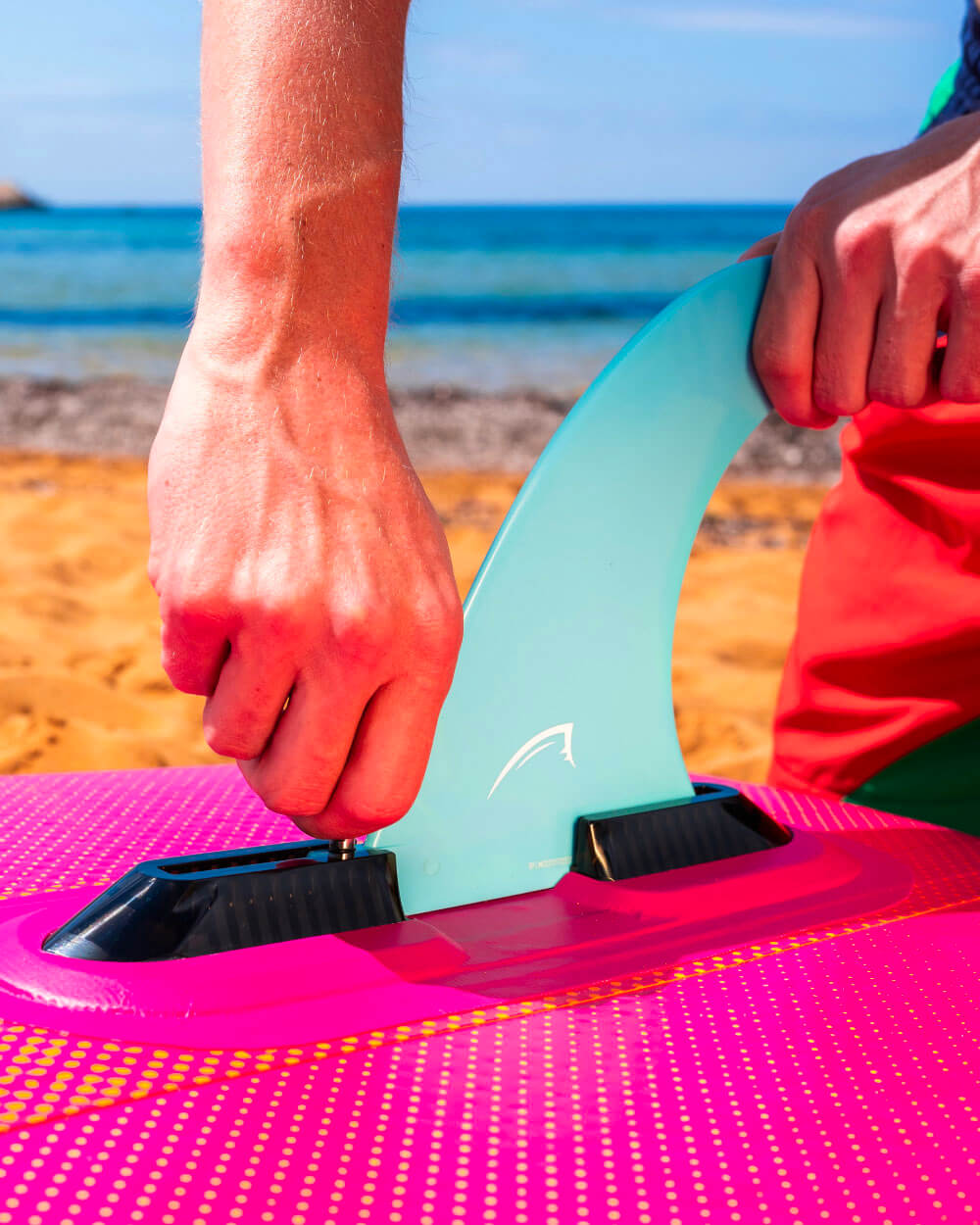 Up close shot of someone setting up the fin on a Bluefin Pink Coral 10' Lite inflatable paddle board on the beach