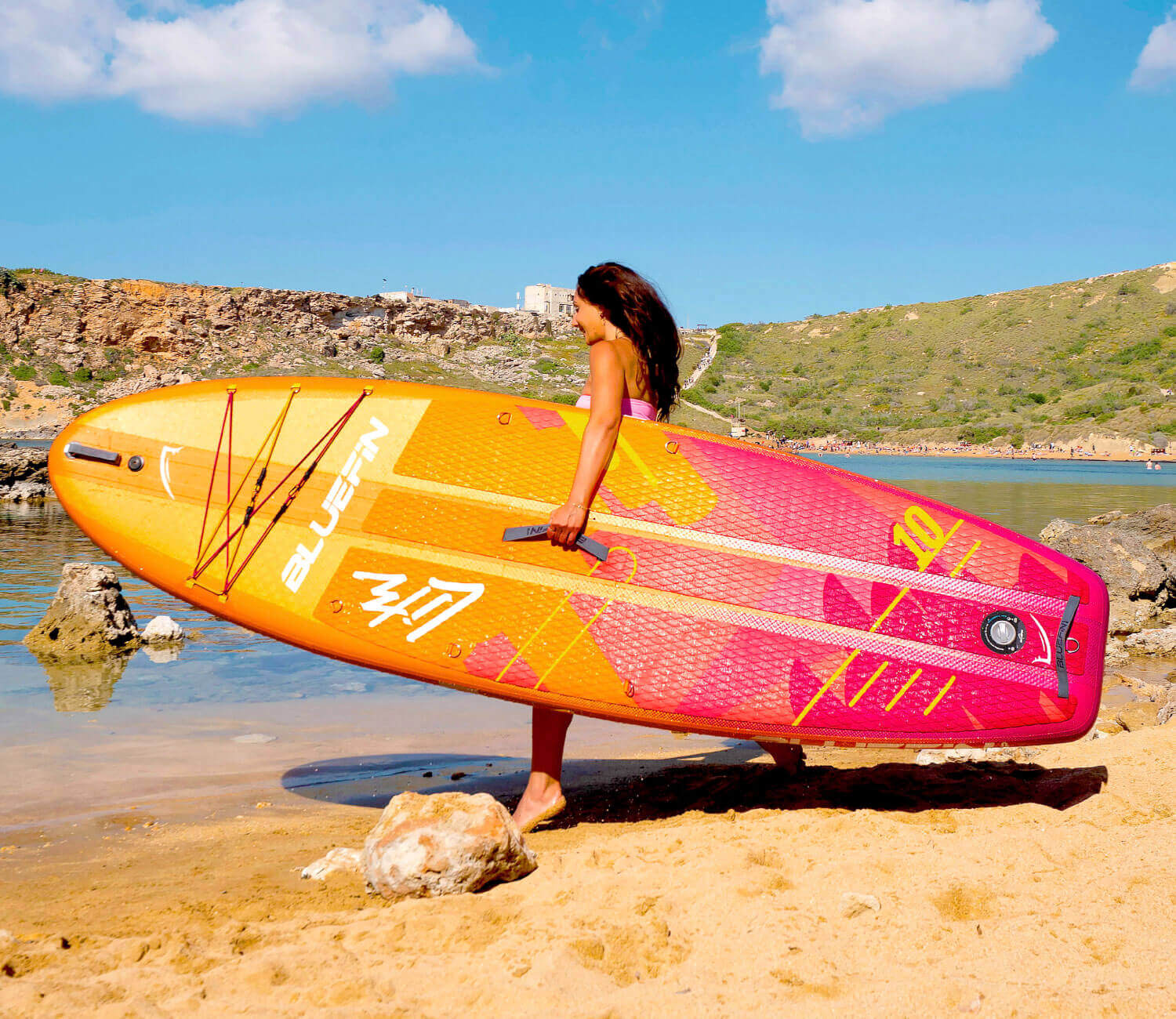 Person carrying a colorful Bluefin paddleboard along a sandy beach near calm water