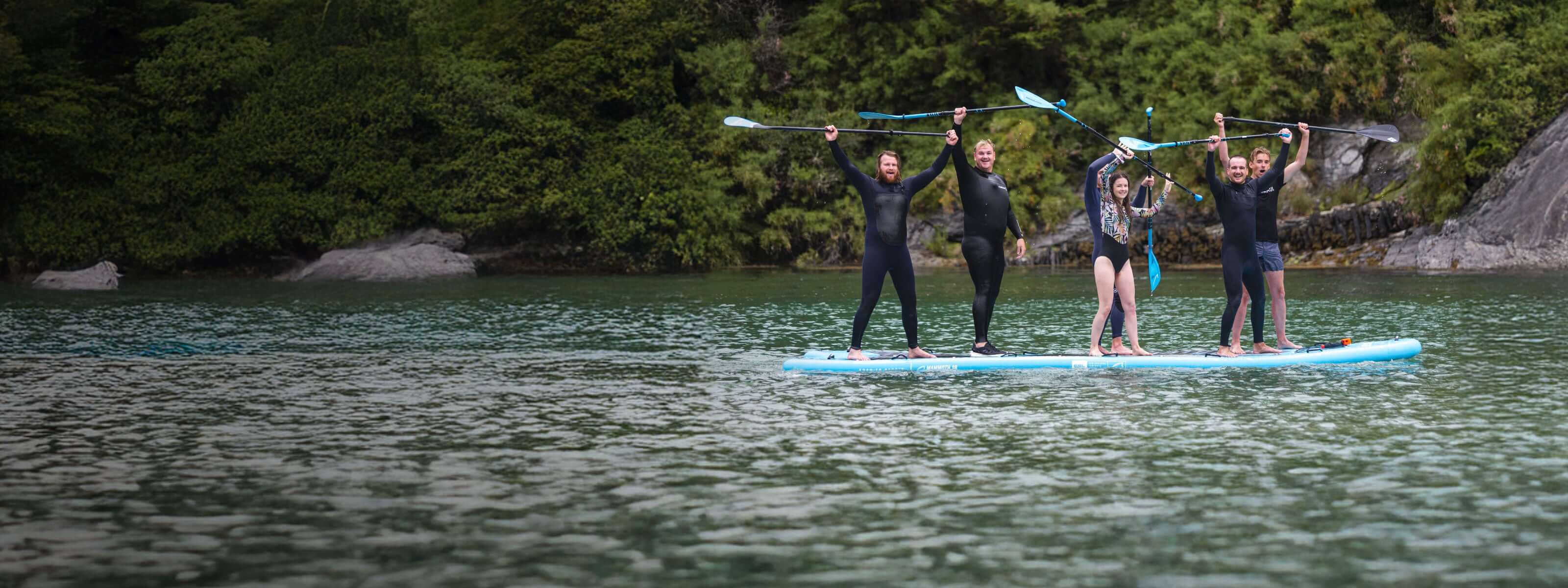 Group of five people on a paddleboard in a serene water setting, celebrating with paddles raised.