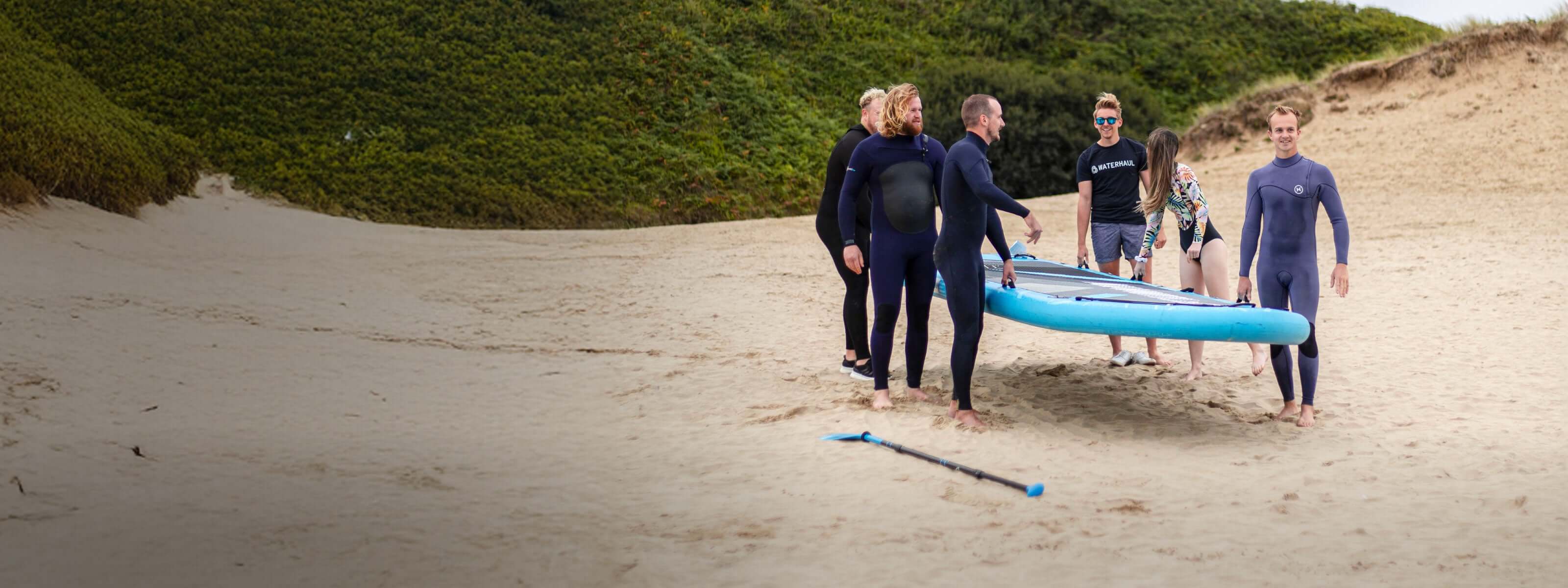 Group of six people in wetsuits carrying a stand-up paddleboard on a sandy beach.