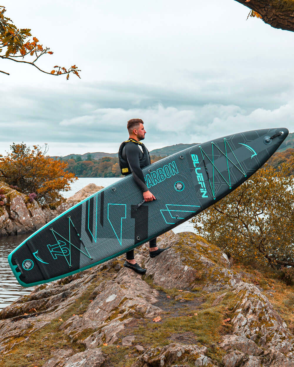 Man in wetsuit carrying a Bluefin Carbon paddleboard near a lake with rocky shore and autumn foliage.