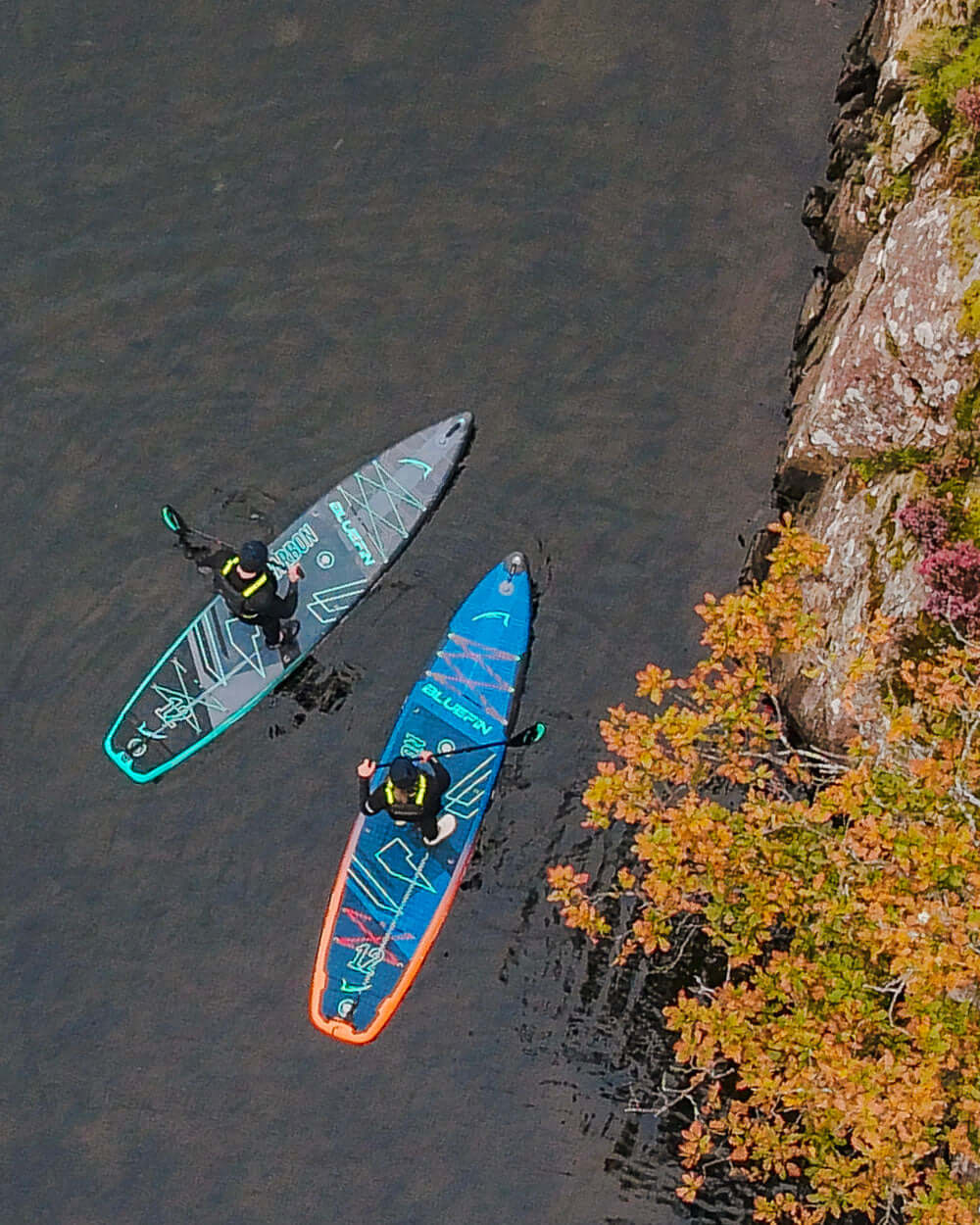 Two paddleboarders on calm water, one in a black and teal board, the other in blue with orange trim.