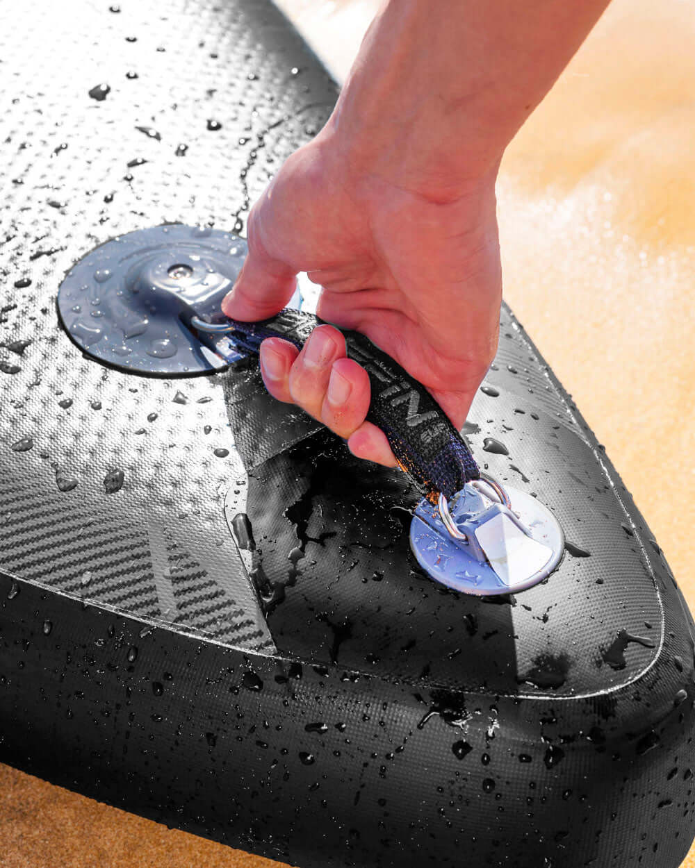 Hand gripping the handle of a wet inflatable paddleboard on a sandy surface