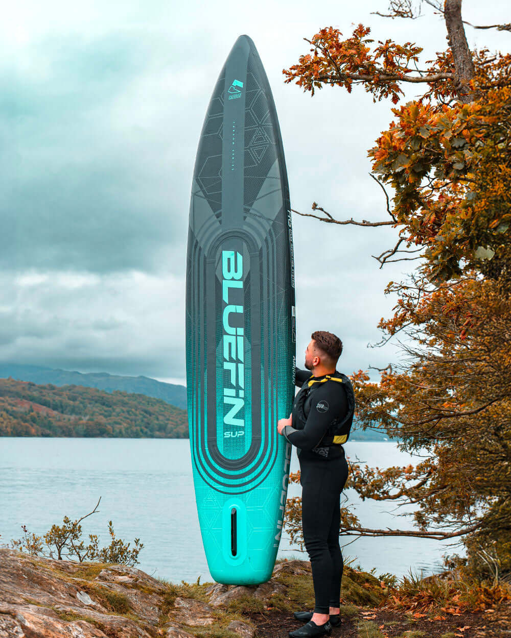 Person in wetsuit beside a Bluefin inflatable paddleboard on a rocky shore by a lake