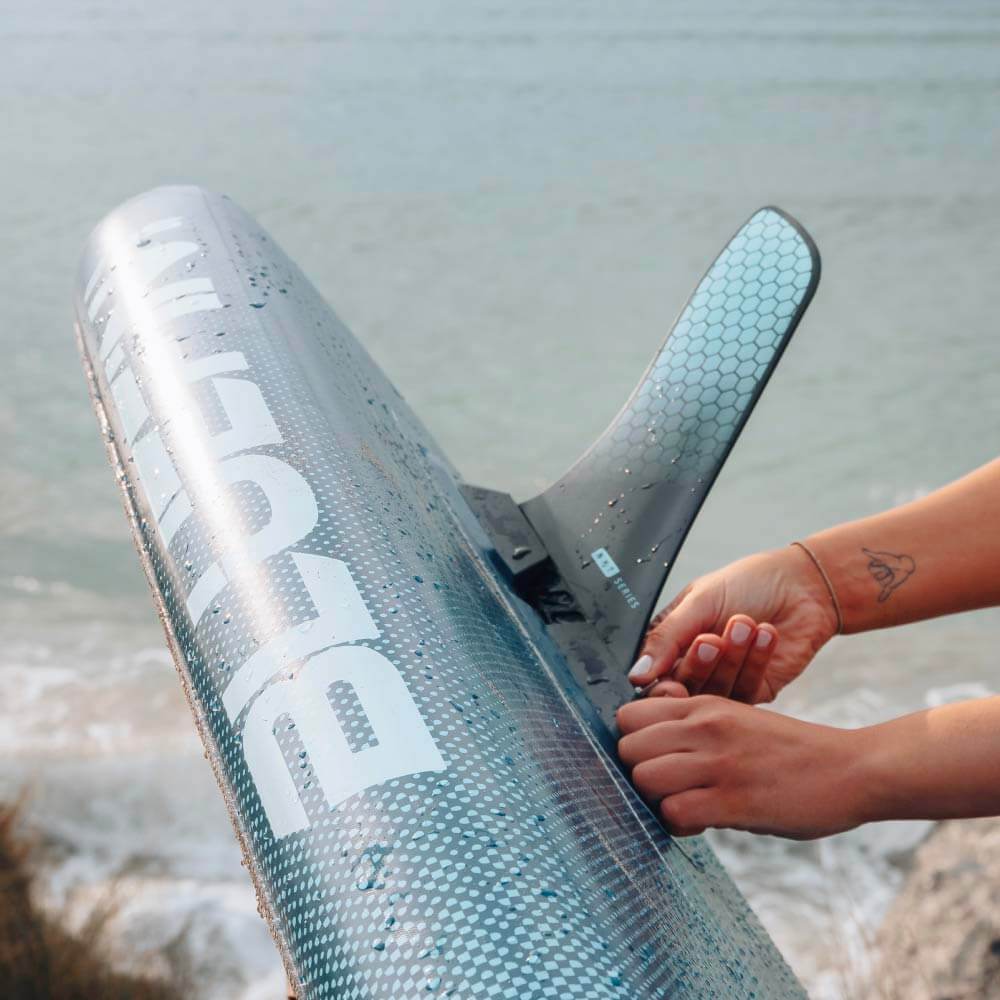 Person attaching a fin to a Bluefin SUP paddleboard by the water's edge.