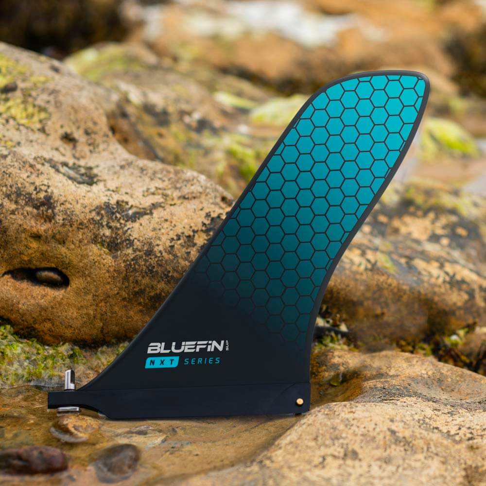 Bluefin NXT Series paddle board fin with teal honeycomb design on rocky surface.