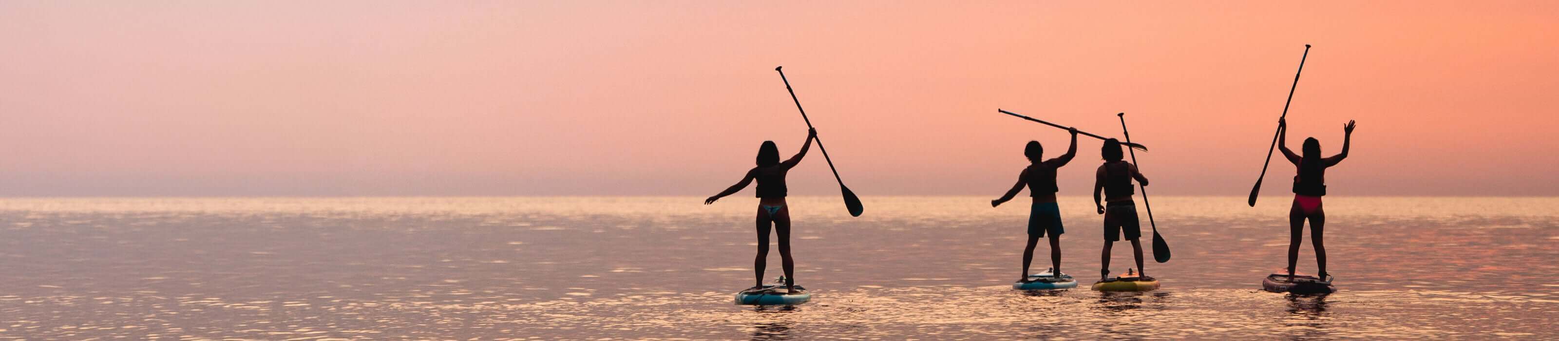 Four people paddleboarding at sunset, silhouetted against a colorful sky on calm water.