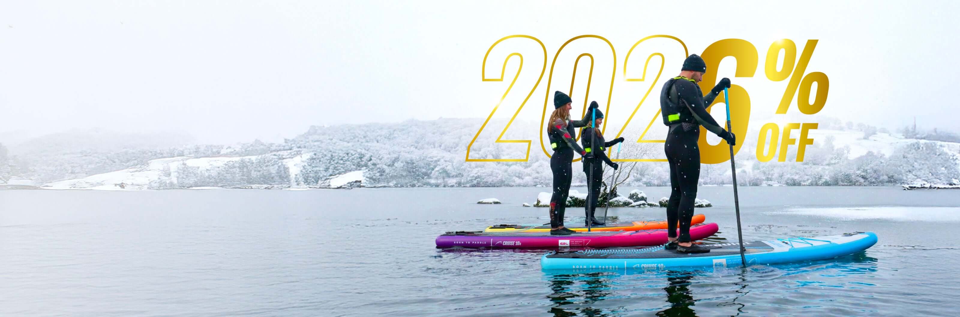 Three paddleboarders on colorful boards in winter scenery with snow-covered hills and water