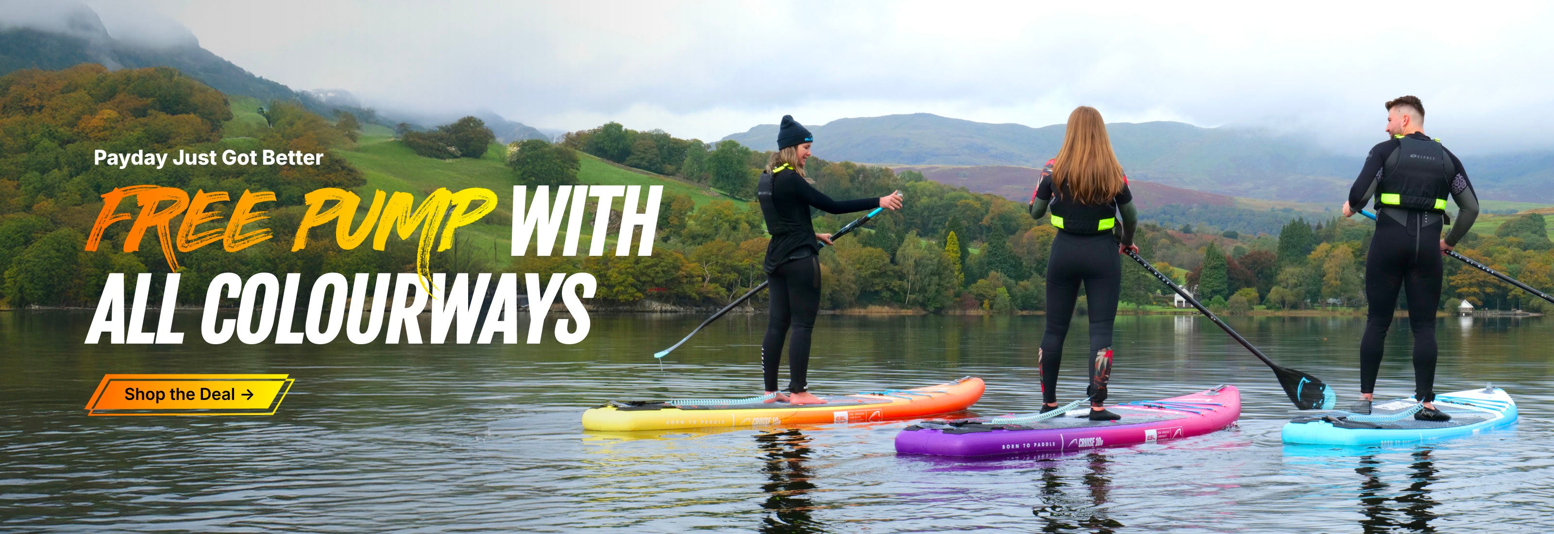 Three people paddleboarding on a lake, wearing wetsuits, with colorful boards in orange, pink, and blue.