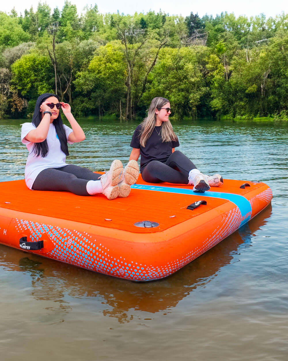 Two people sitting on an inflatable play air dock in a body of water with trees in the background