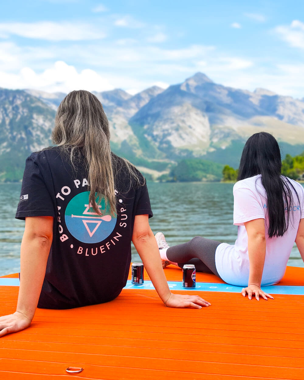 Two people sitting on a play air dock with mountains and water in the background