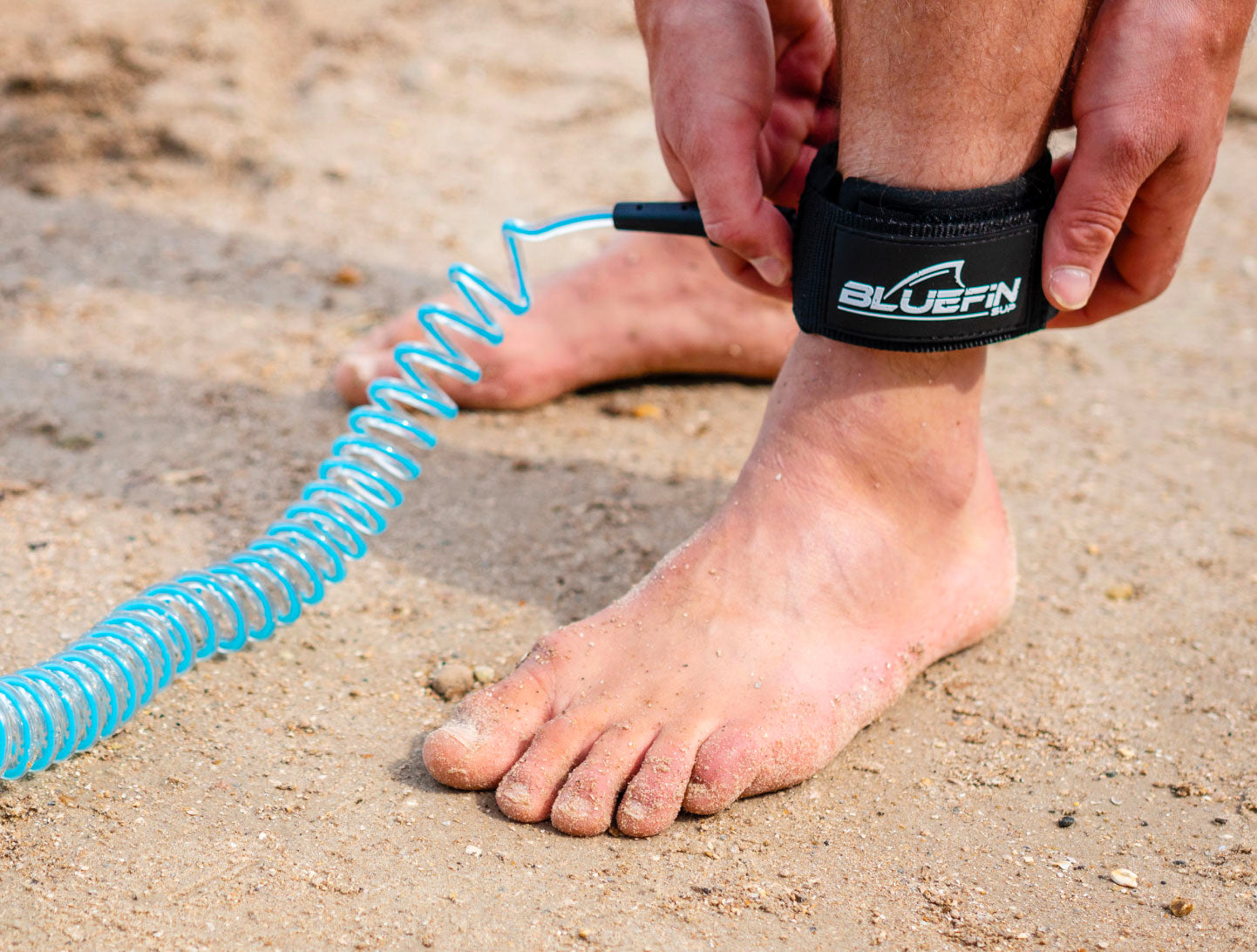 Person securing a Bluefin SUP ankle leash on sandy beach.