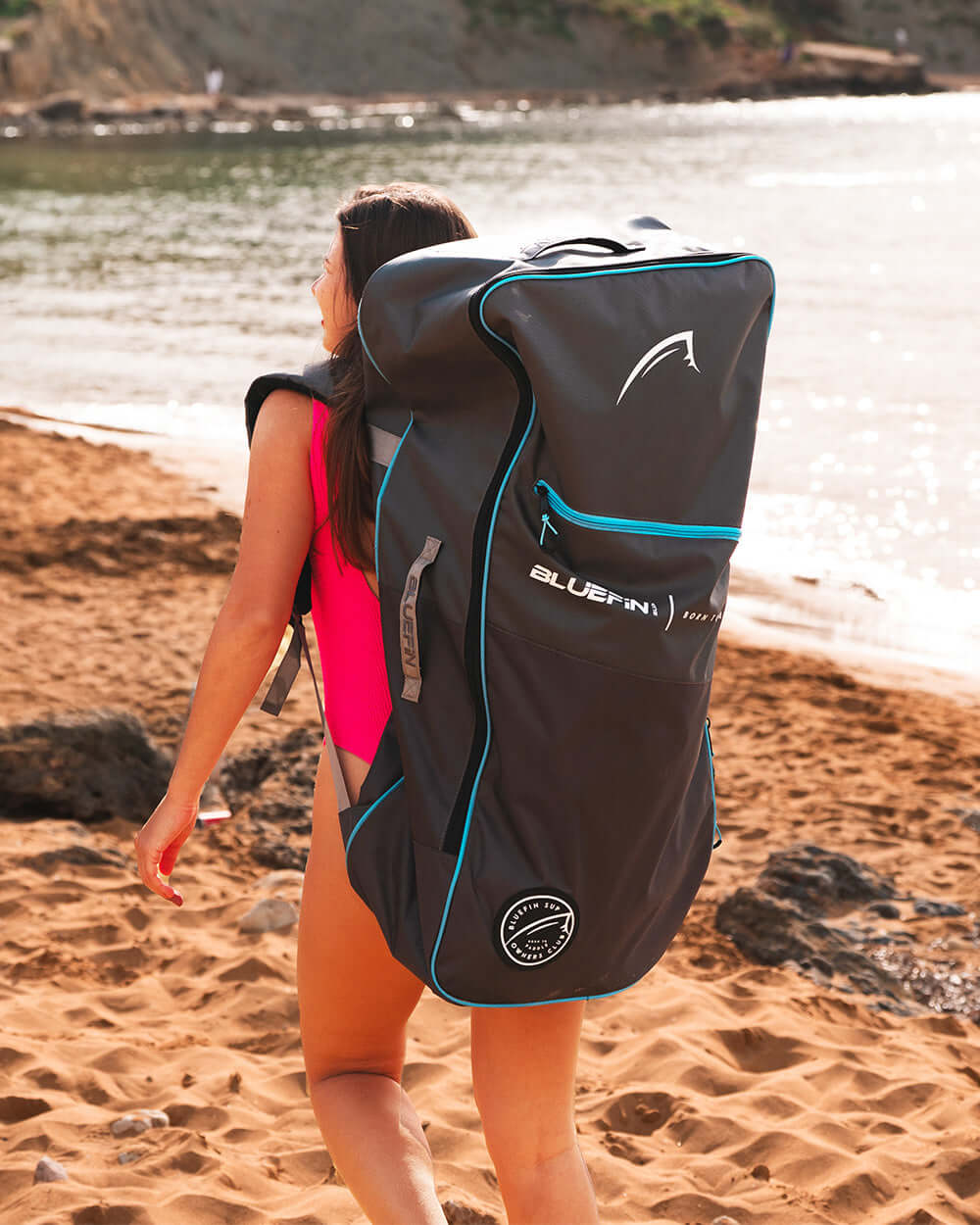 Person in a pink swimsuit carrying a large Bluefin SUP backpack on a sandy beach near water