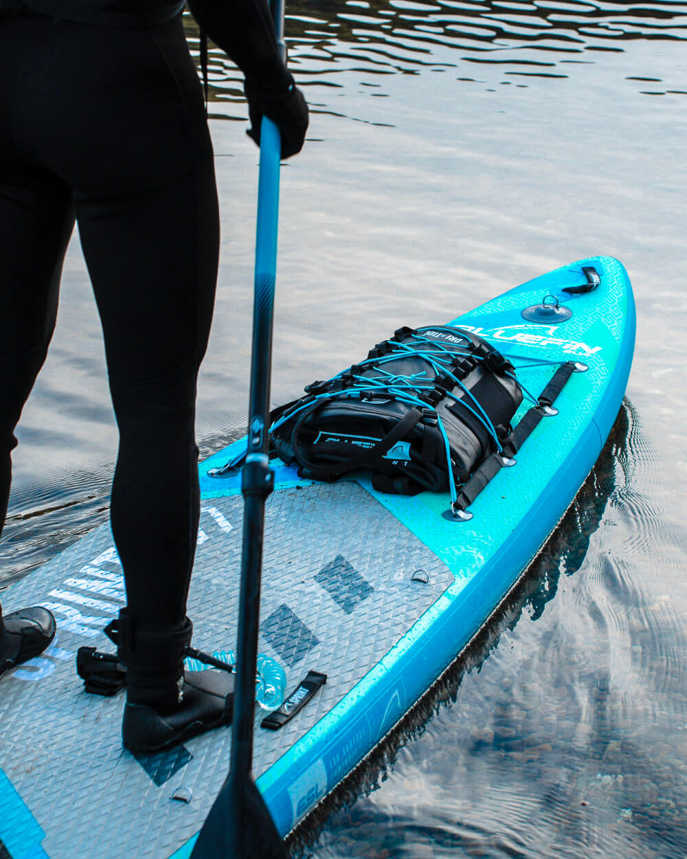 Person on a blue sprint paddleboard with a backpack underneath the bungee cords standing in water.