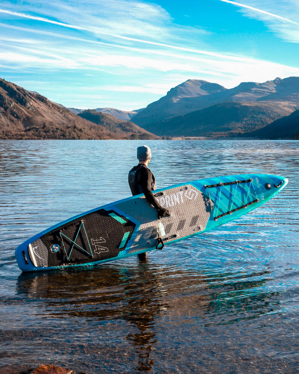 Person holding a blue and black sprint paddleboard on a calm lake with mountains in the background