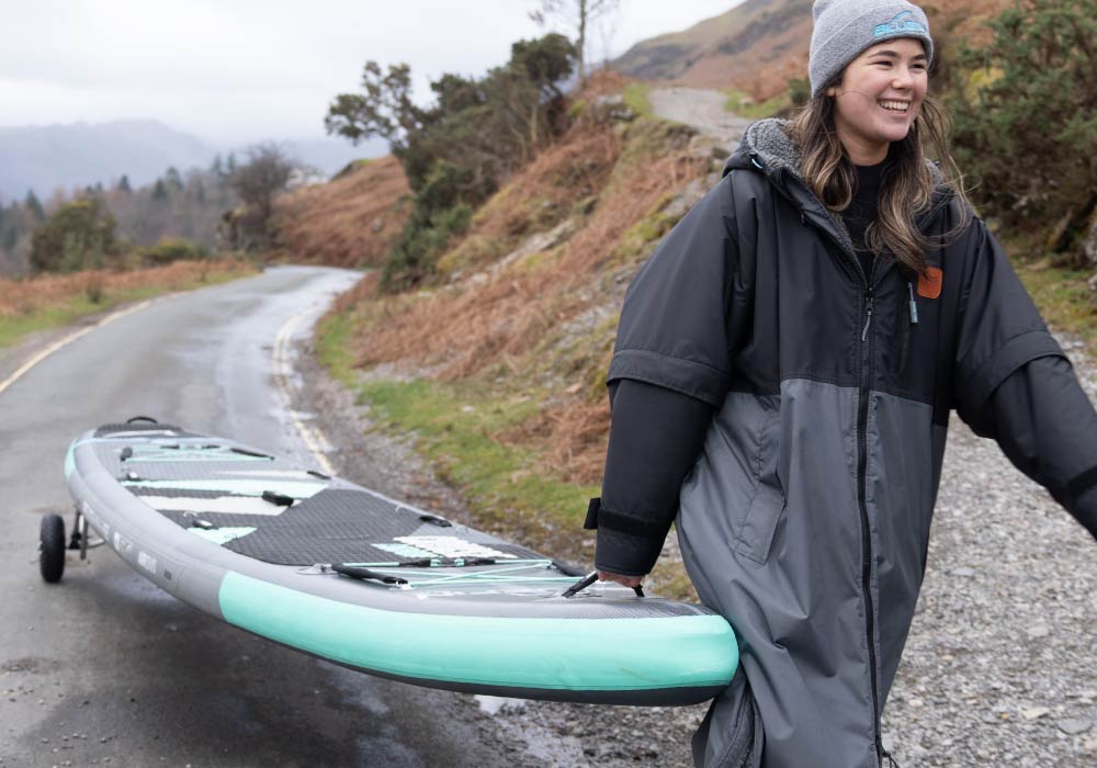Smiling individual carries a paddleboard along a scenic road, wearing a cozy jacket and beanie.