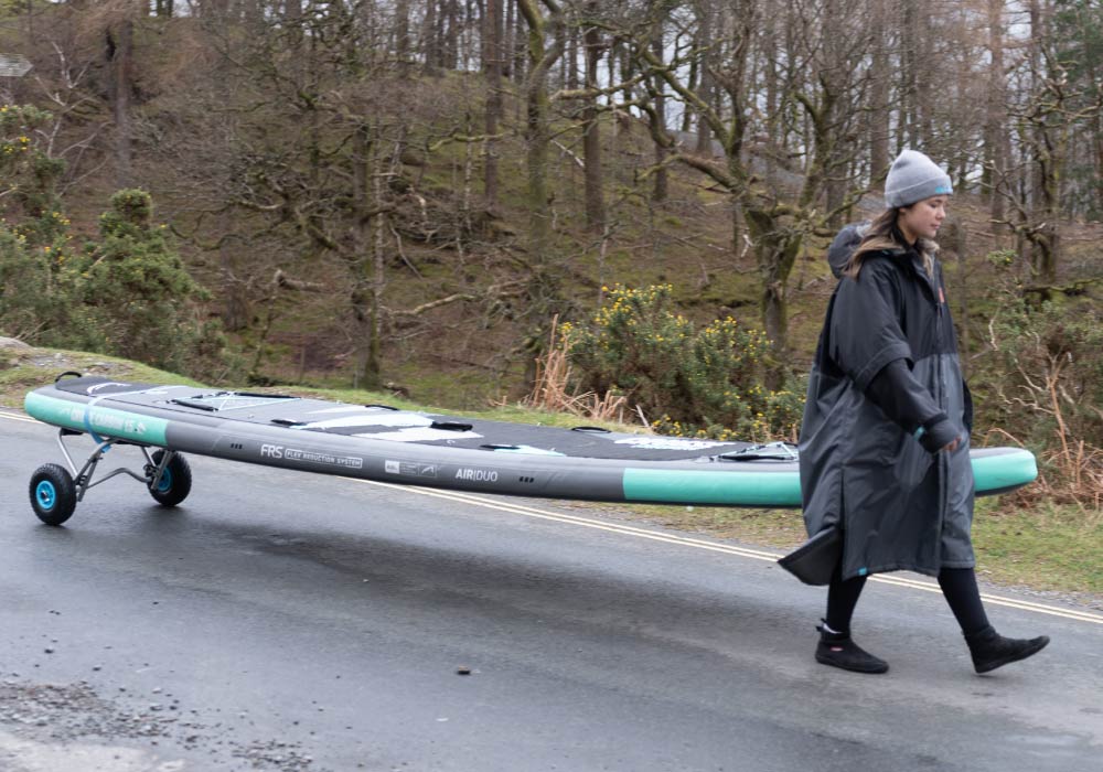 Person walking with a paddleboard on wheels along a road in a wooded area.