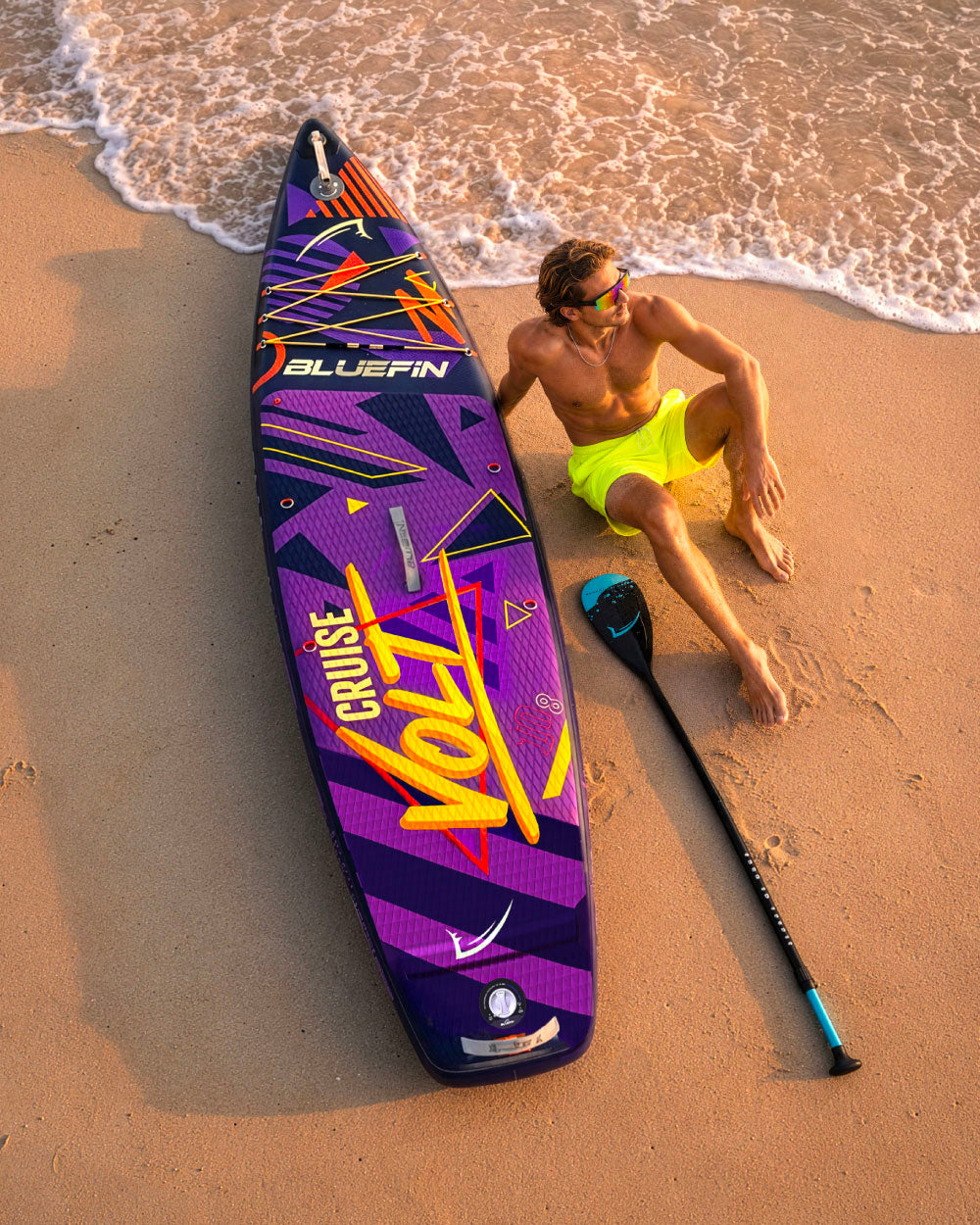 Inflatable paddleboard with colorful design, man in yellow shorts relaxing on sand, paddle nearby
