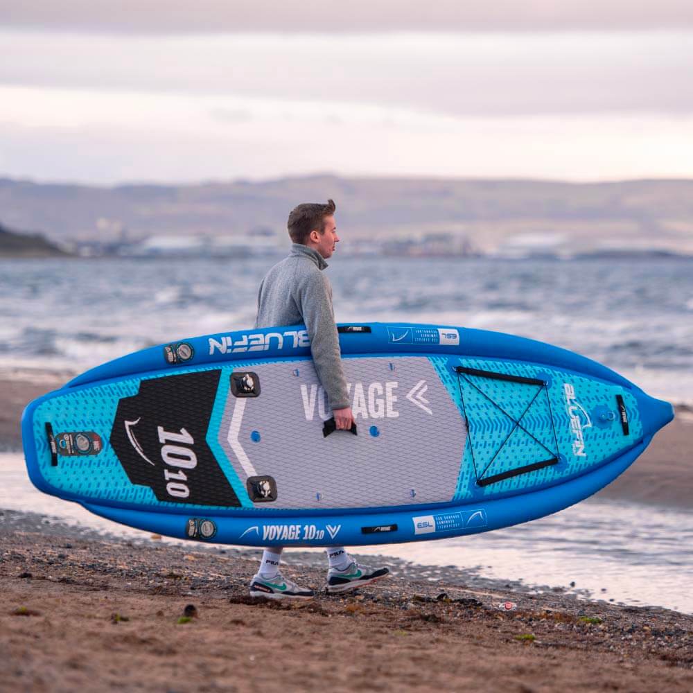 Person carrying a blue paddleboard on the beach with water in the background.