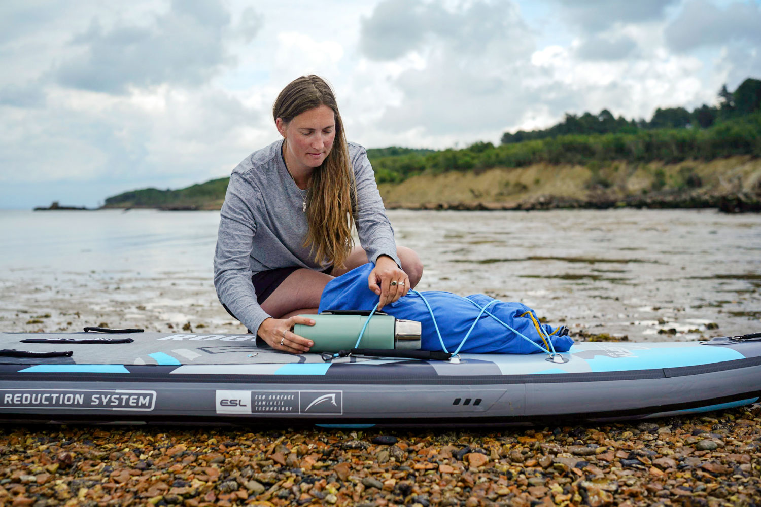 Woman packing gear on an inflatable stand-up paddleboard by the shore, preparing for an adventure.