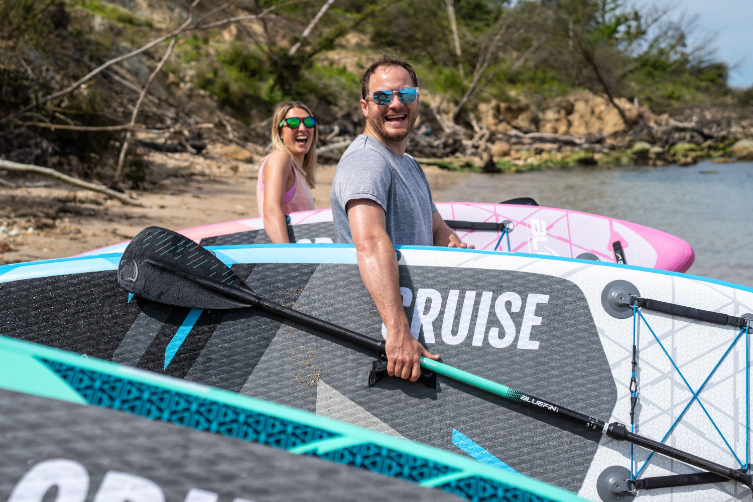 Two joyful friends at the beach, ready for paddleboarding with colorful boards and paddles.