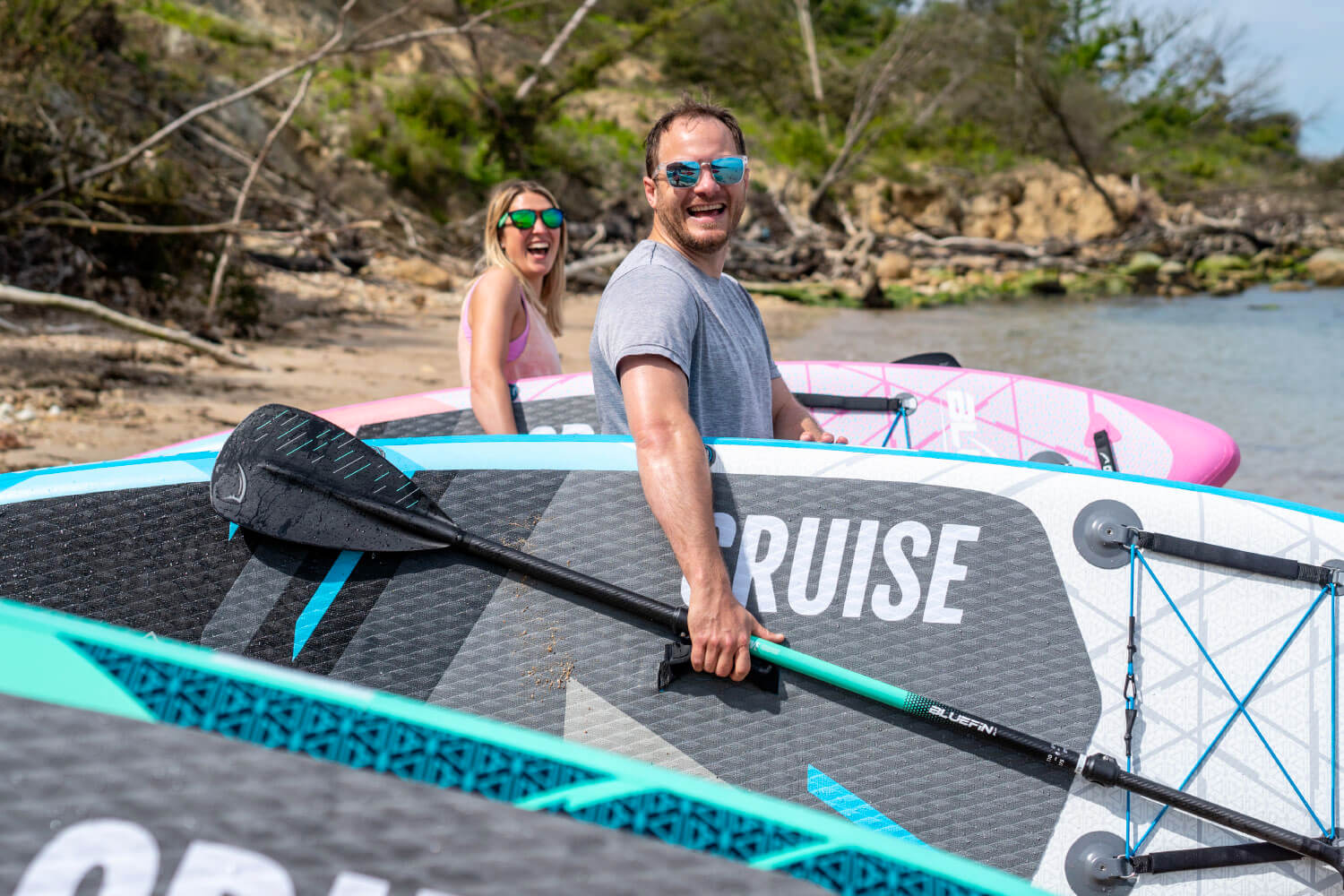 Two joyful friends at the beach, ready for paddleboarding with colorful boards and paddles.