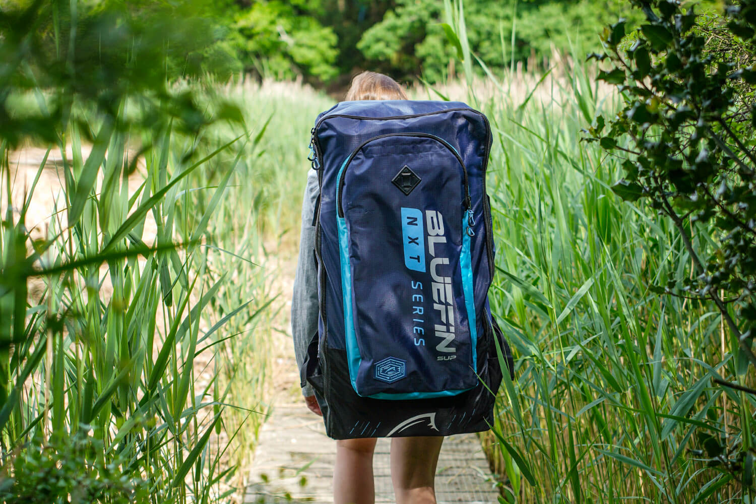 Person carrying a Bluefin SUP backpack through a grassy pathway. Ideal for outdoor adventures.