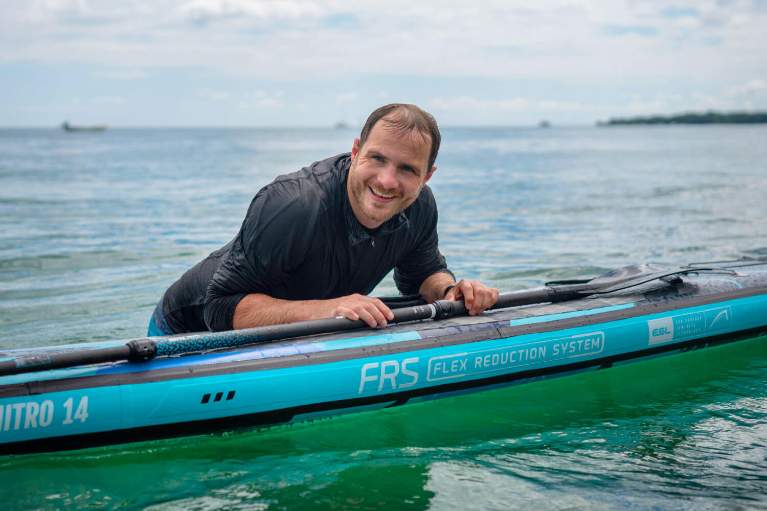 Smiling man leaning on a Bluefin SUP board in clear water, showcasing the FRS Flex Reduction System.