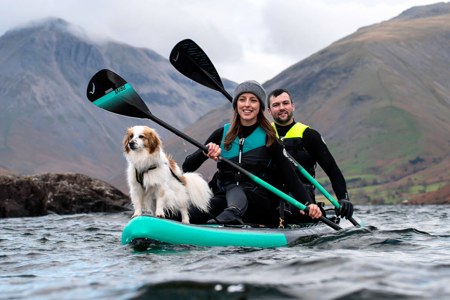 Two people and a dog enjoy paddleboarding on a lake with mountains in the background.