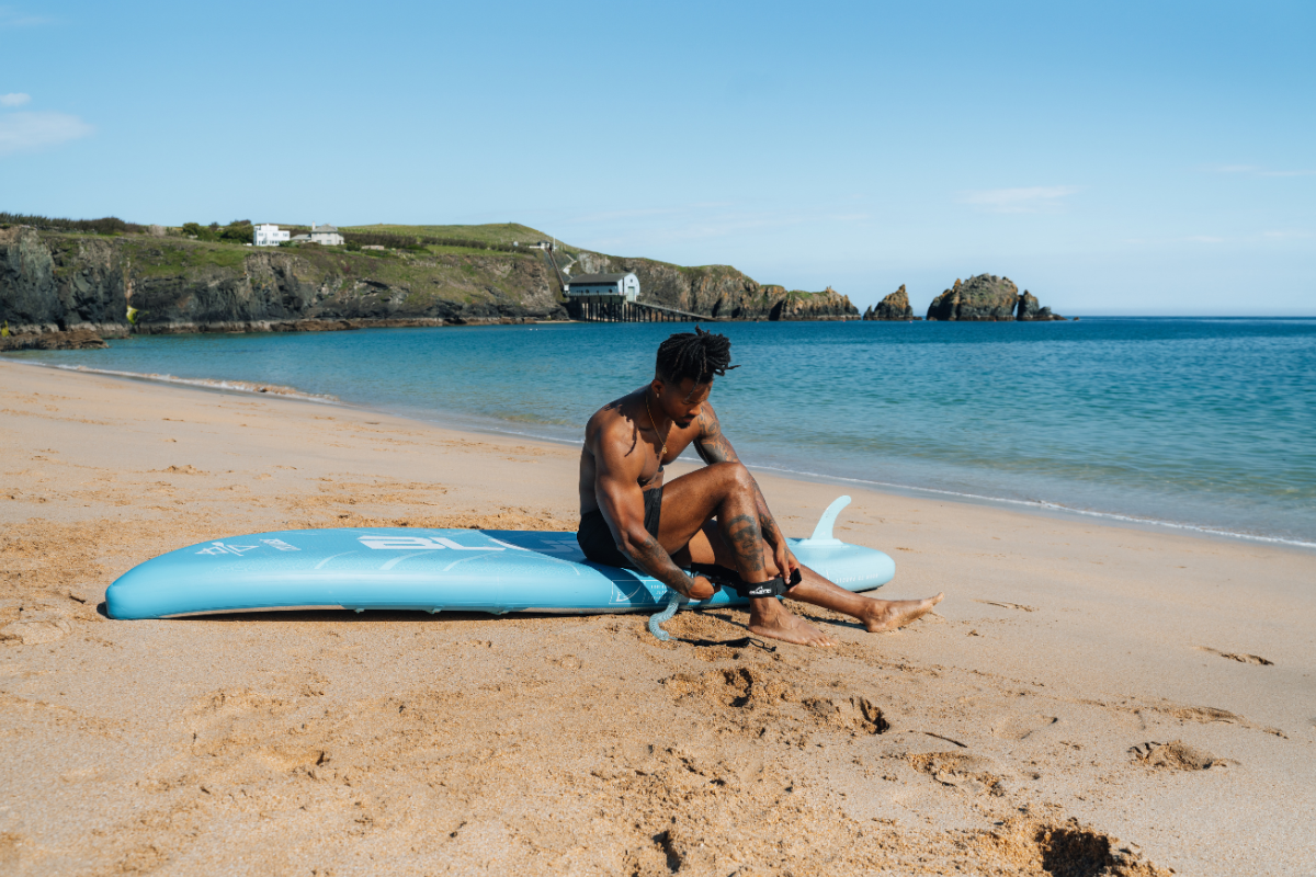man sat on a paddleboard on the sand attaching his ankle leash