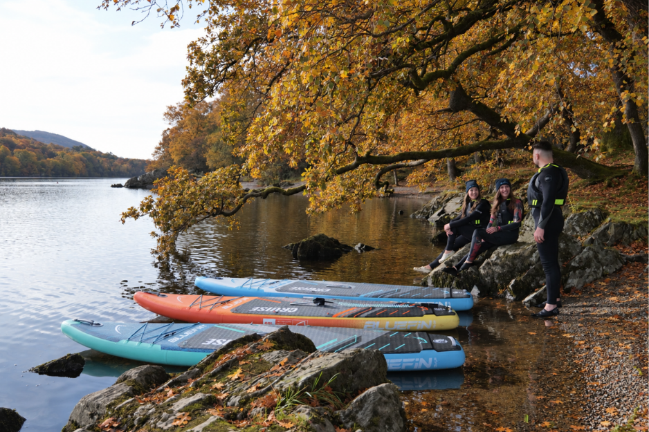 three people sat by an autumnal lake with their paddleboards pointing towards the water