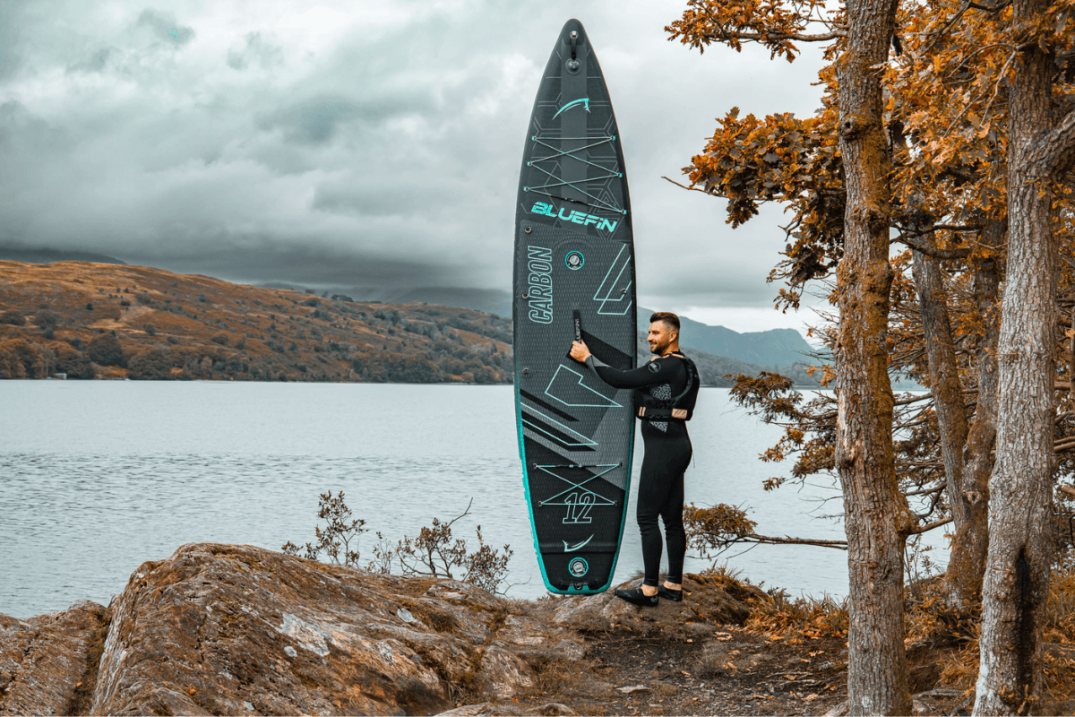 man holding a paddleboard by the edge of a lake 